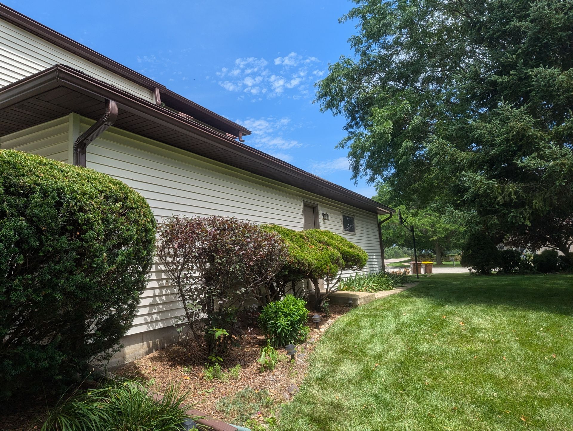 Beige house exterior with brown trim, green bushes, and blue sky.