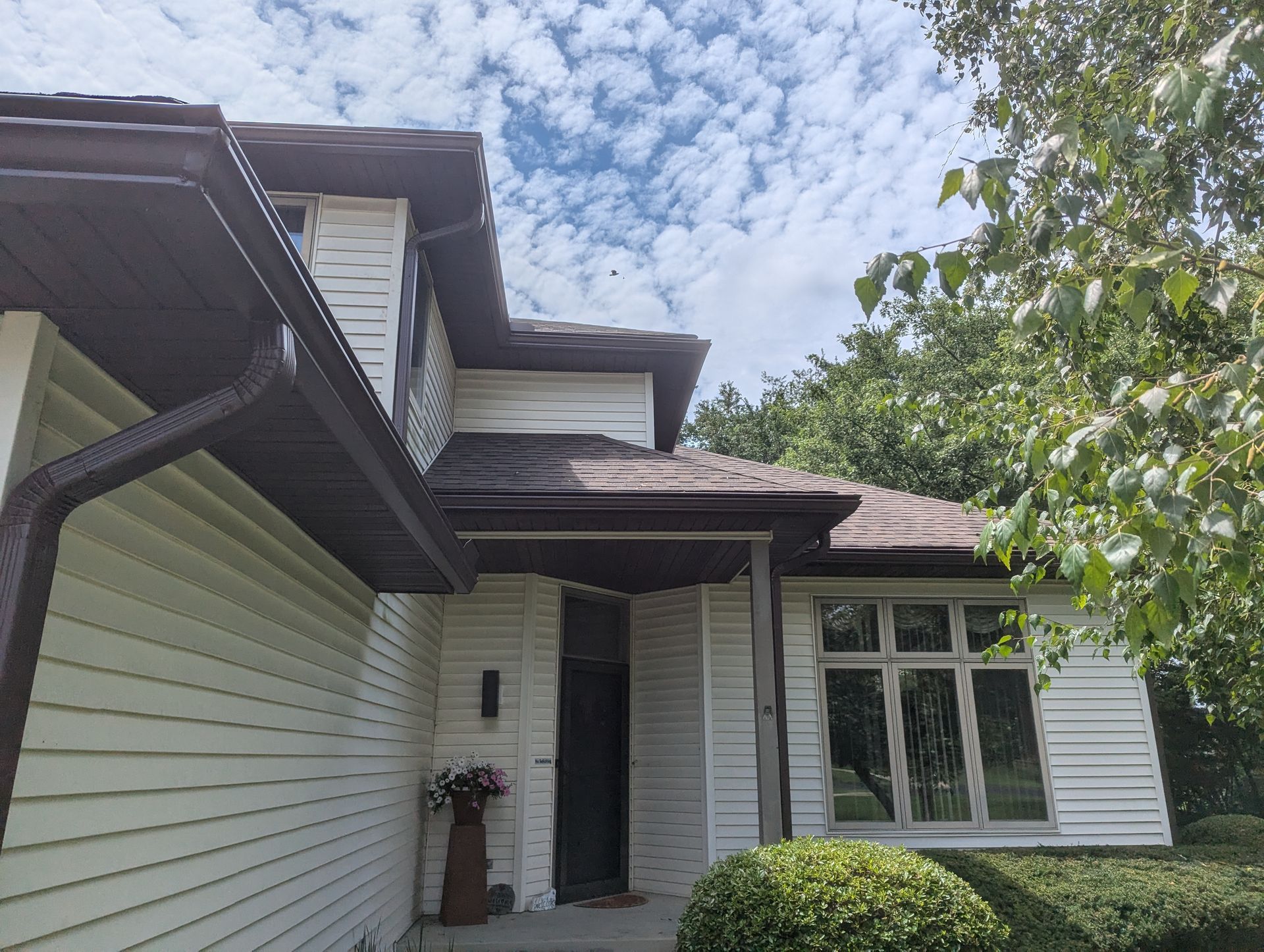 White house with brown trim and gutters, under a blue sky with clouds, surrounded by greenery.