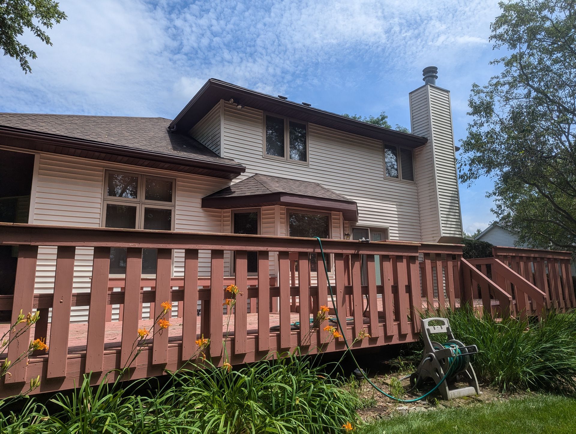 Deck with a red railing, beige siding on a two-story house, and a brick chimney under a cloudy sky.