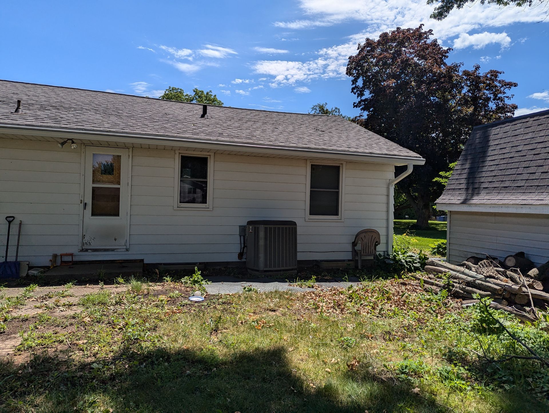 Back of a white house with a brown roof and a small building to the right, overgrown grass in the foreground.