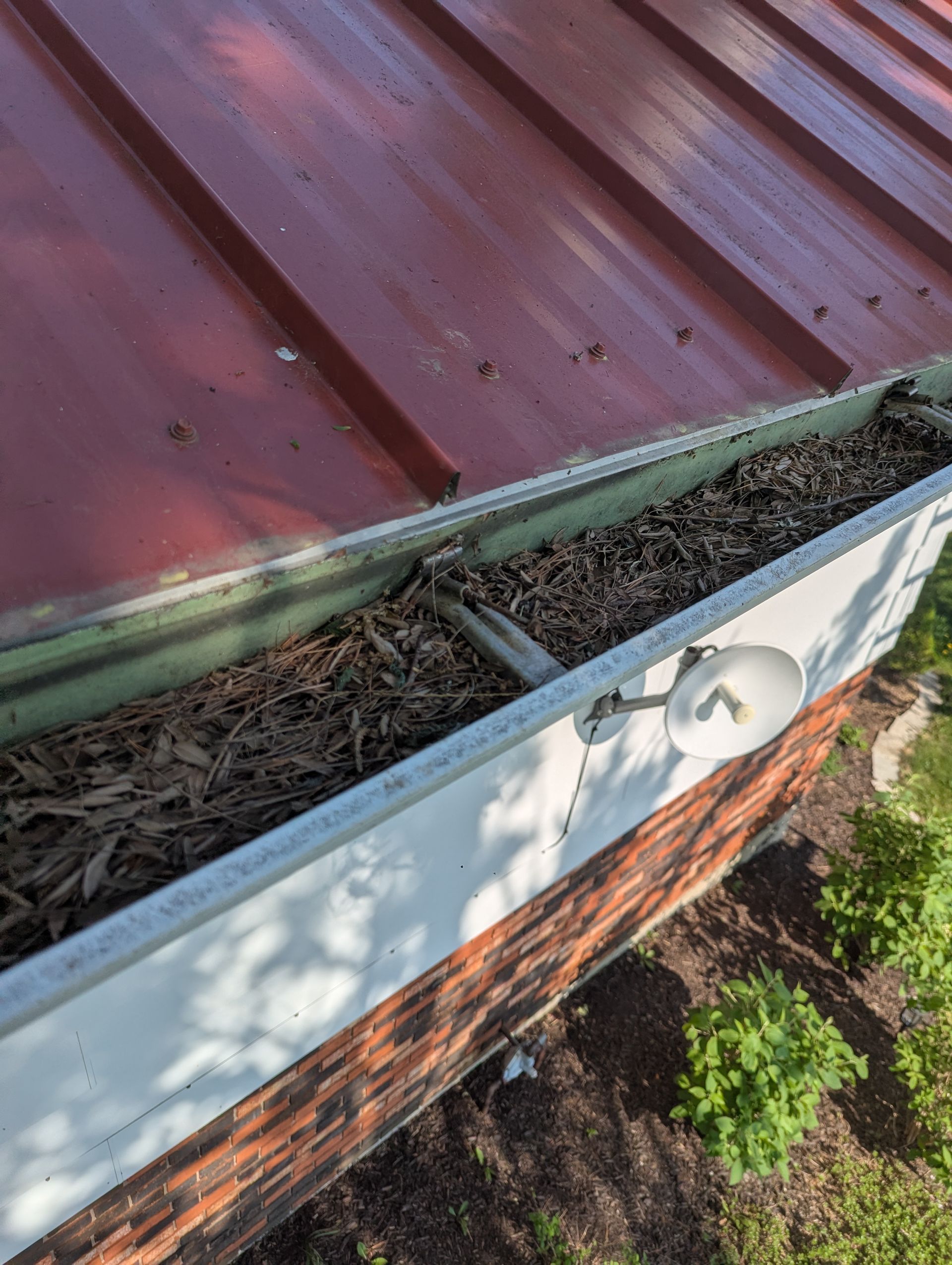 Red roof gutter filled with leaves, next to a brick wall. A white light is attached.
