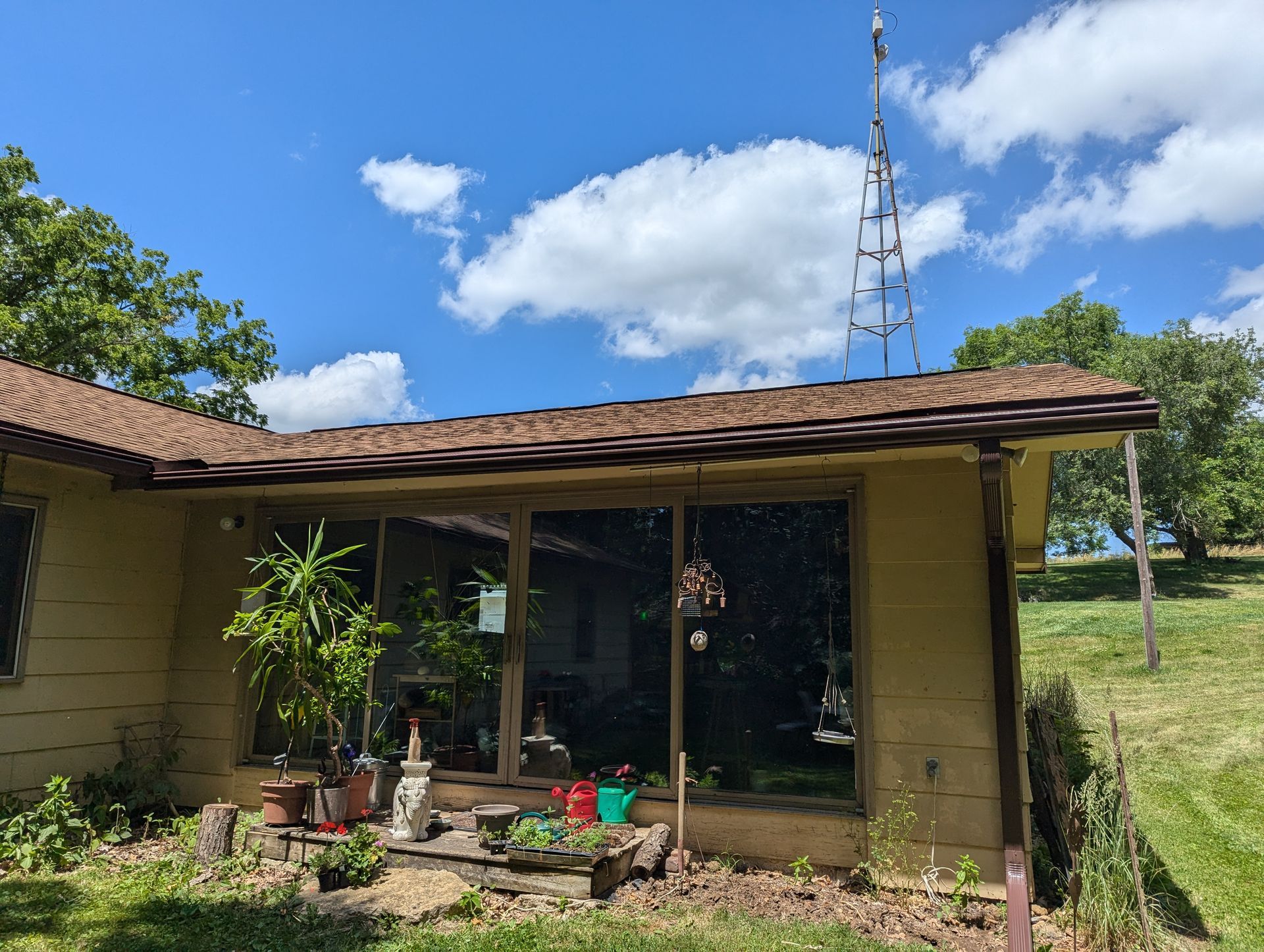 A house with large windows, potted plants, and a radio tower on the roof under a blue sky with fluffy clouds.