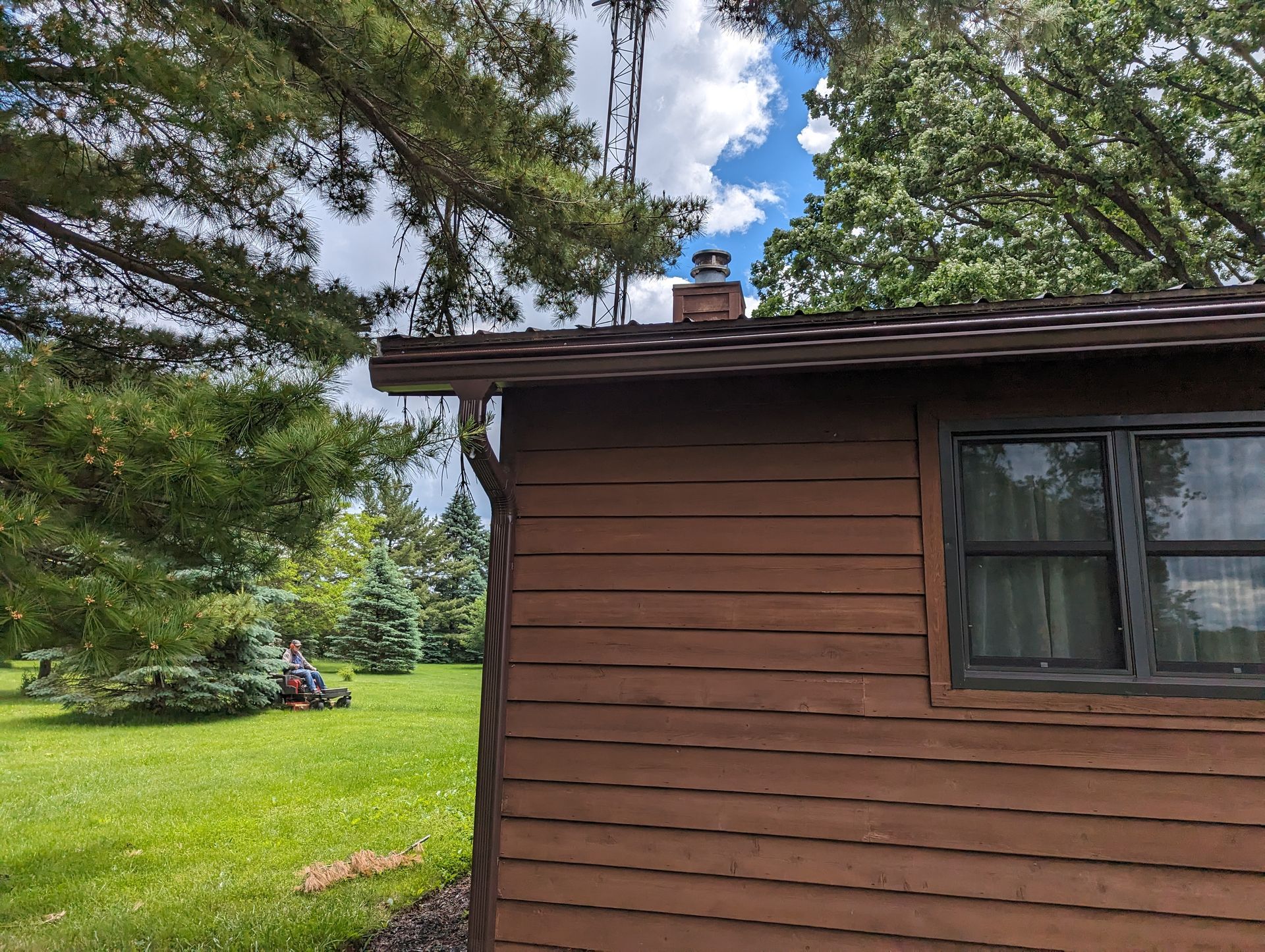 Brown cabin exterior with window, chimney, and green yard under a partly cloudy sky.