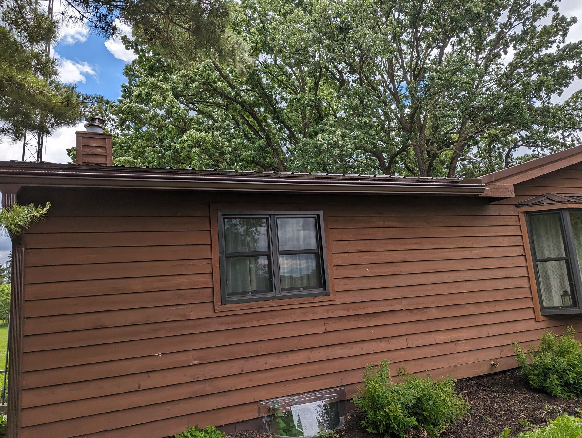 Brown house with dark window frame, brown siding and a rooftop with a chimney. Green foliage in the background.