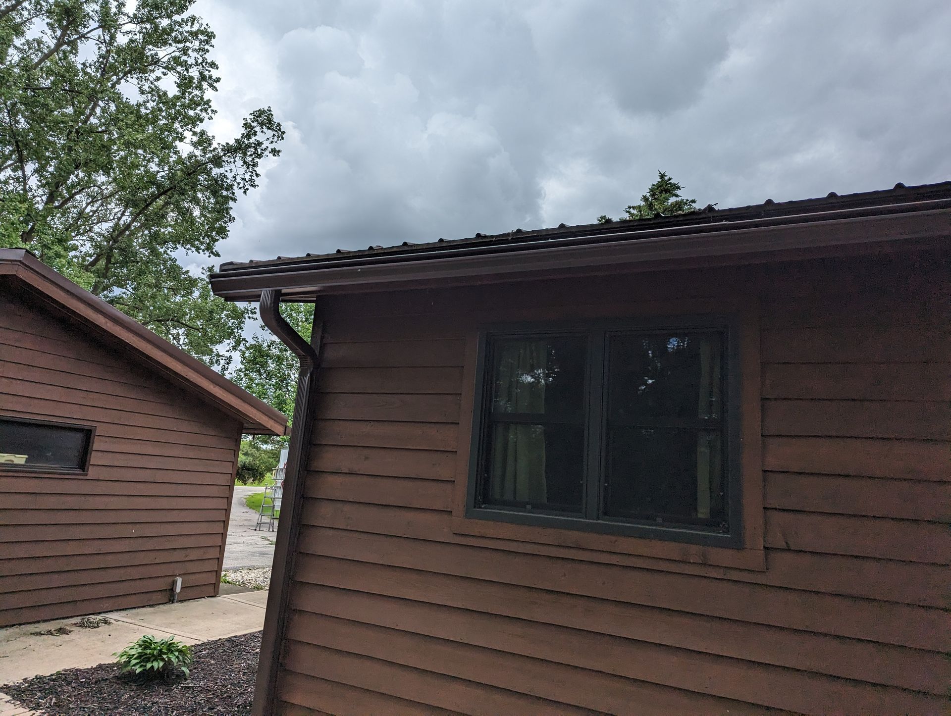 Brown-sided building with dark window and gutter. Cloudy sky and tree in the background.