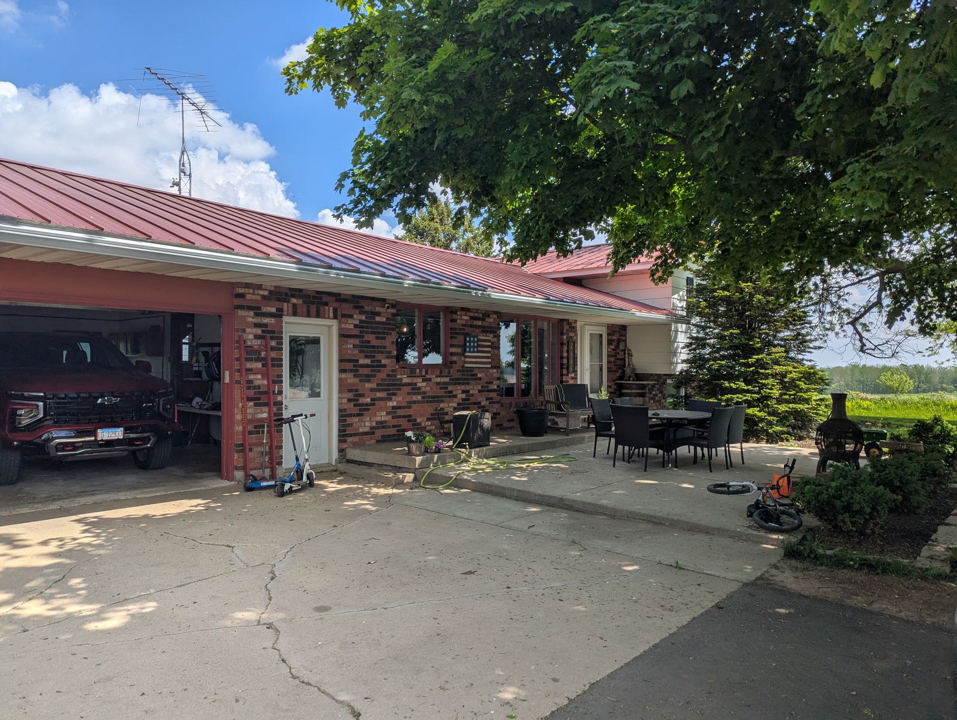 Brick house with red roof, attached garage, and patio under trees on a sunny day.