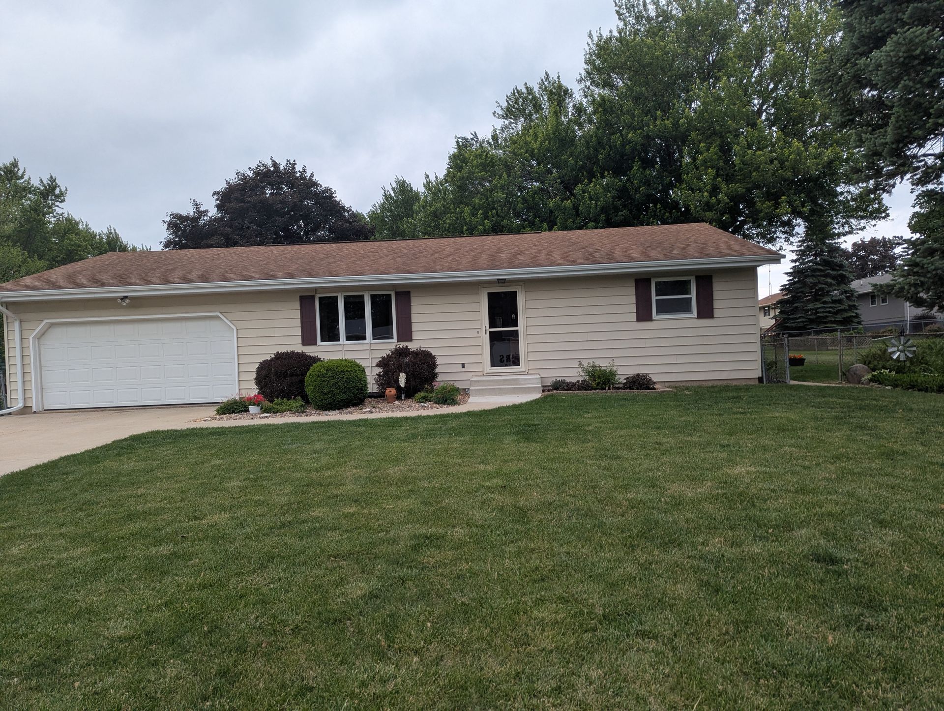 Tan ranch-style house with a brown roof and a white garage door, set in front of a green lawn under a cloudy sky.