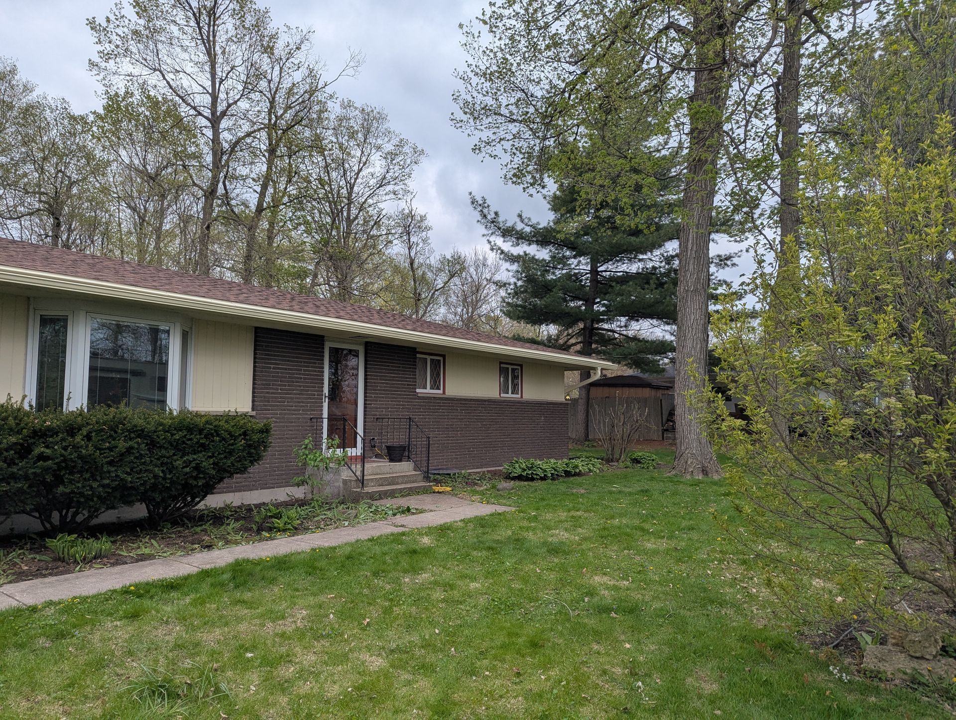 A one-story beige and brick house with green lawn and trees under a cloudy sky.