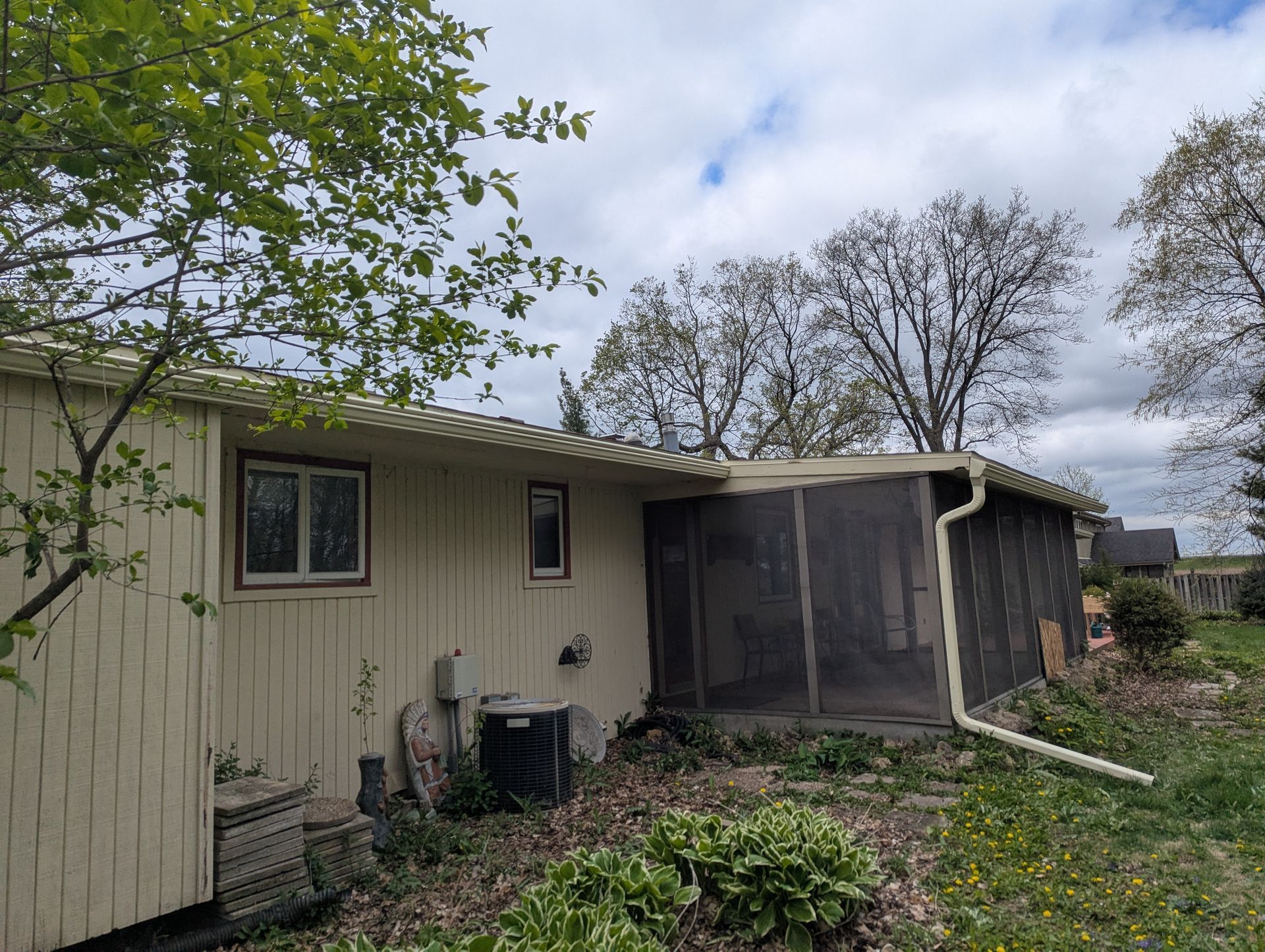 Back of a beige house with a screened porch, windows, and overgrown landscaping under a cloudy sky.
