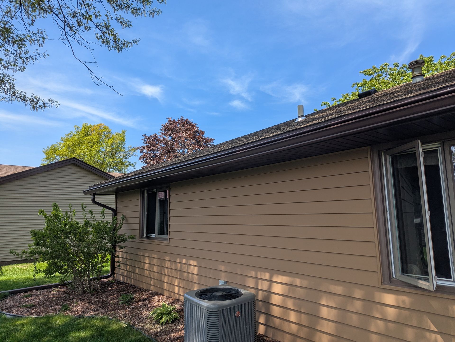 Side view of a brown-sided house with a dark brown roof and gutter. Two windows are open. Blue sky.