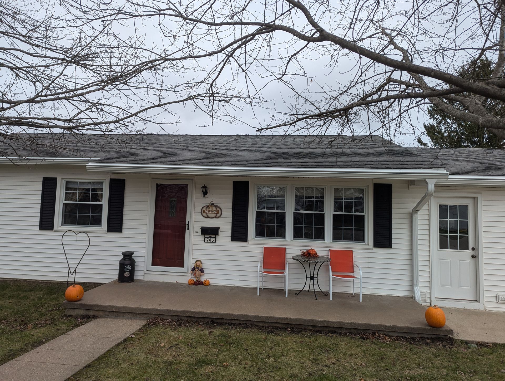 White house with black shutters, red door, pumpkins, and two orange chairs on porch.