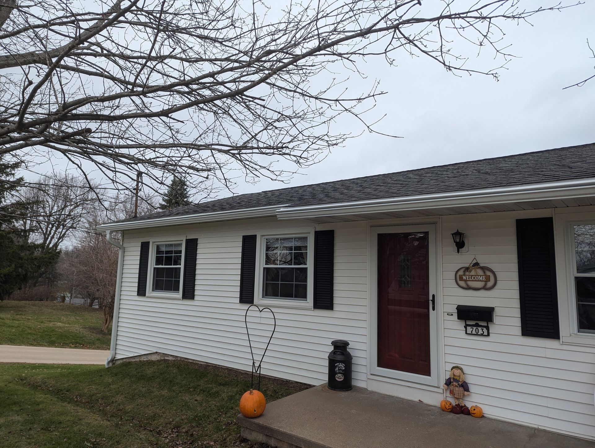 White house with black shutters, a red door, and fall decorations under a cloudy sky.