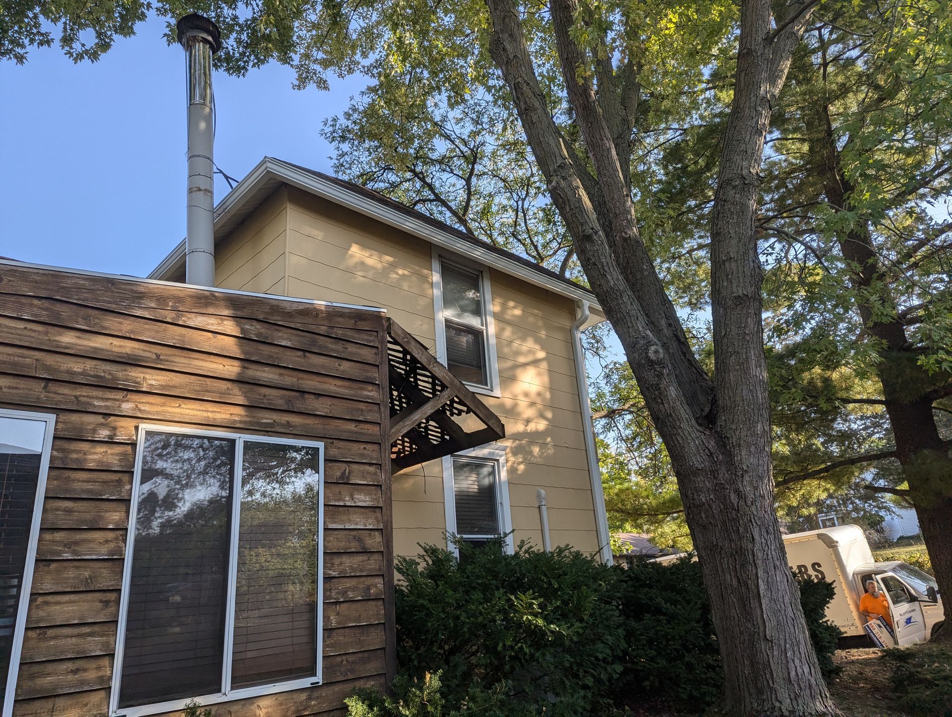 Two-story yellow house with a chimney, awning, and a brown log-sided structure to the left.