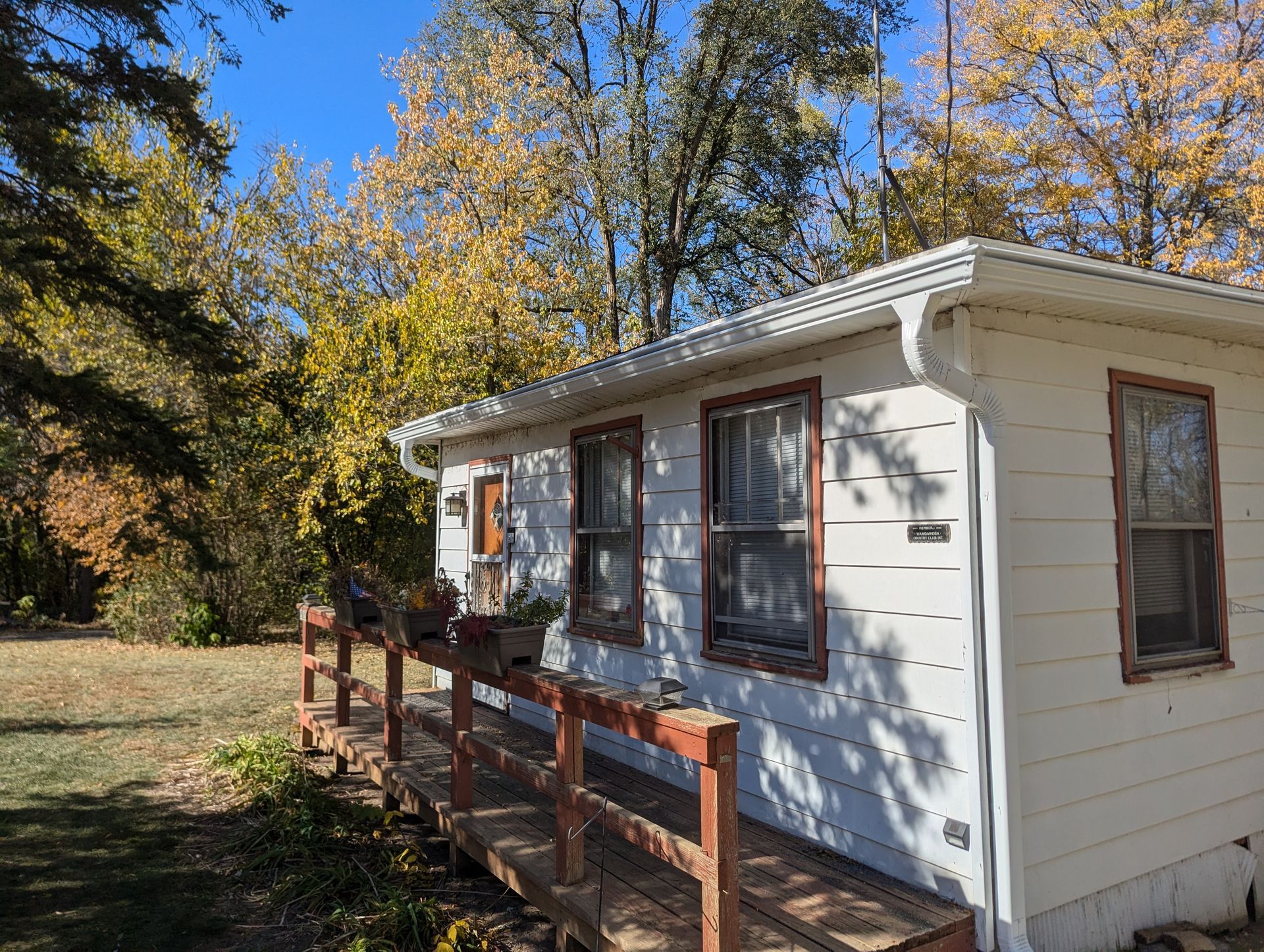 White one-story house with a wooden ramp; surrounded by trees with autumn foliage under a blue sky.