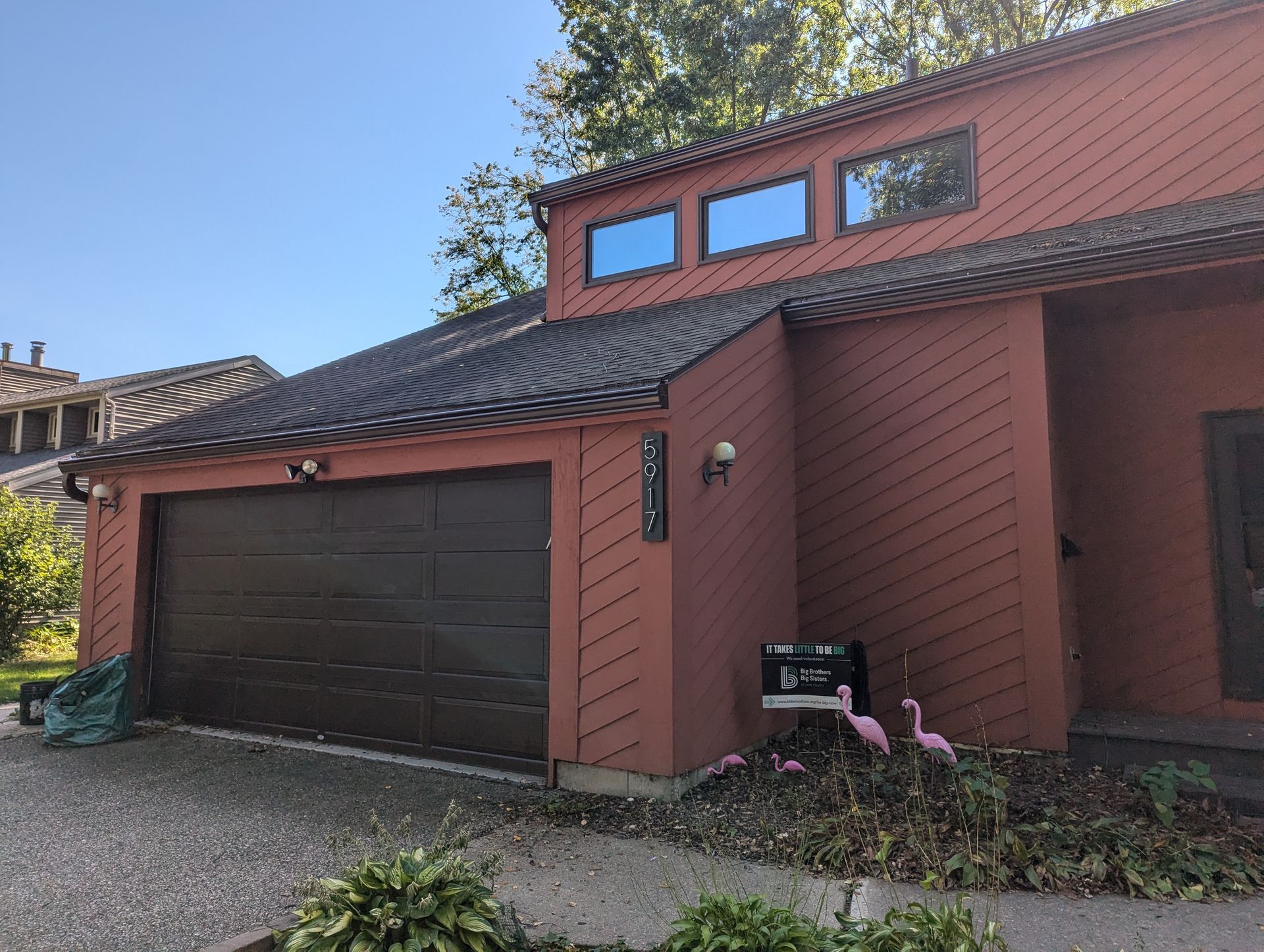 Brown house with a garage and three windows on the roof. Pink flamingos in the yard.