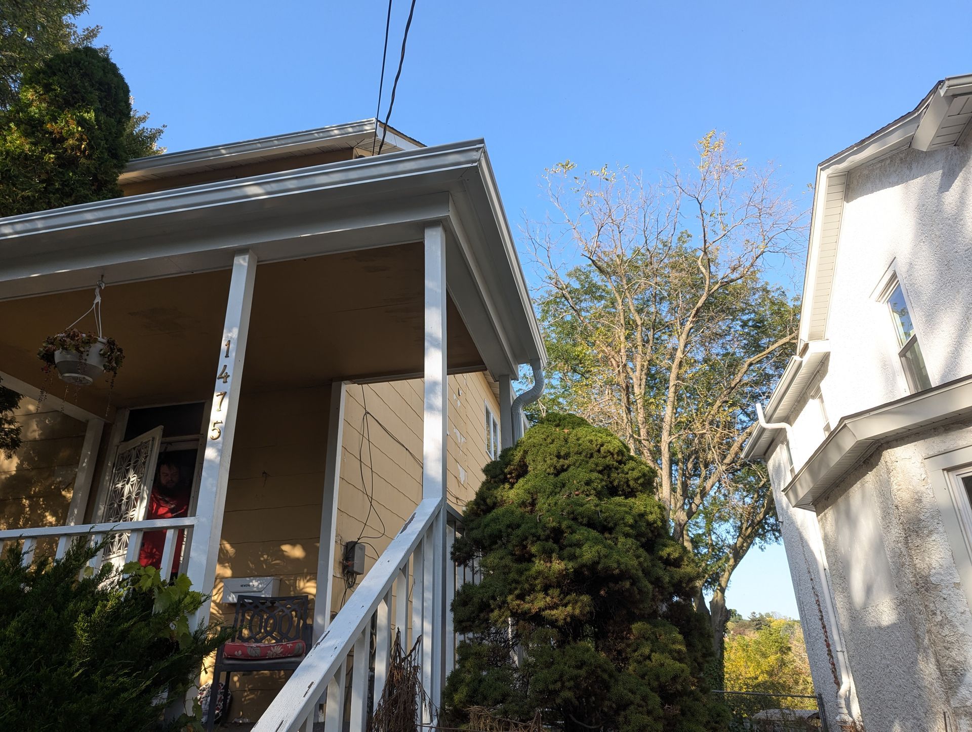 Yellow house with white trim and porch, next to a white house with trees and clear sky.