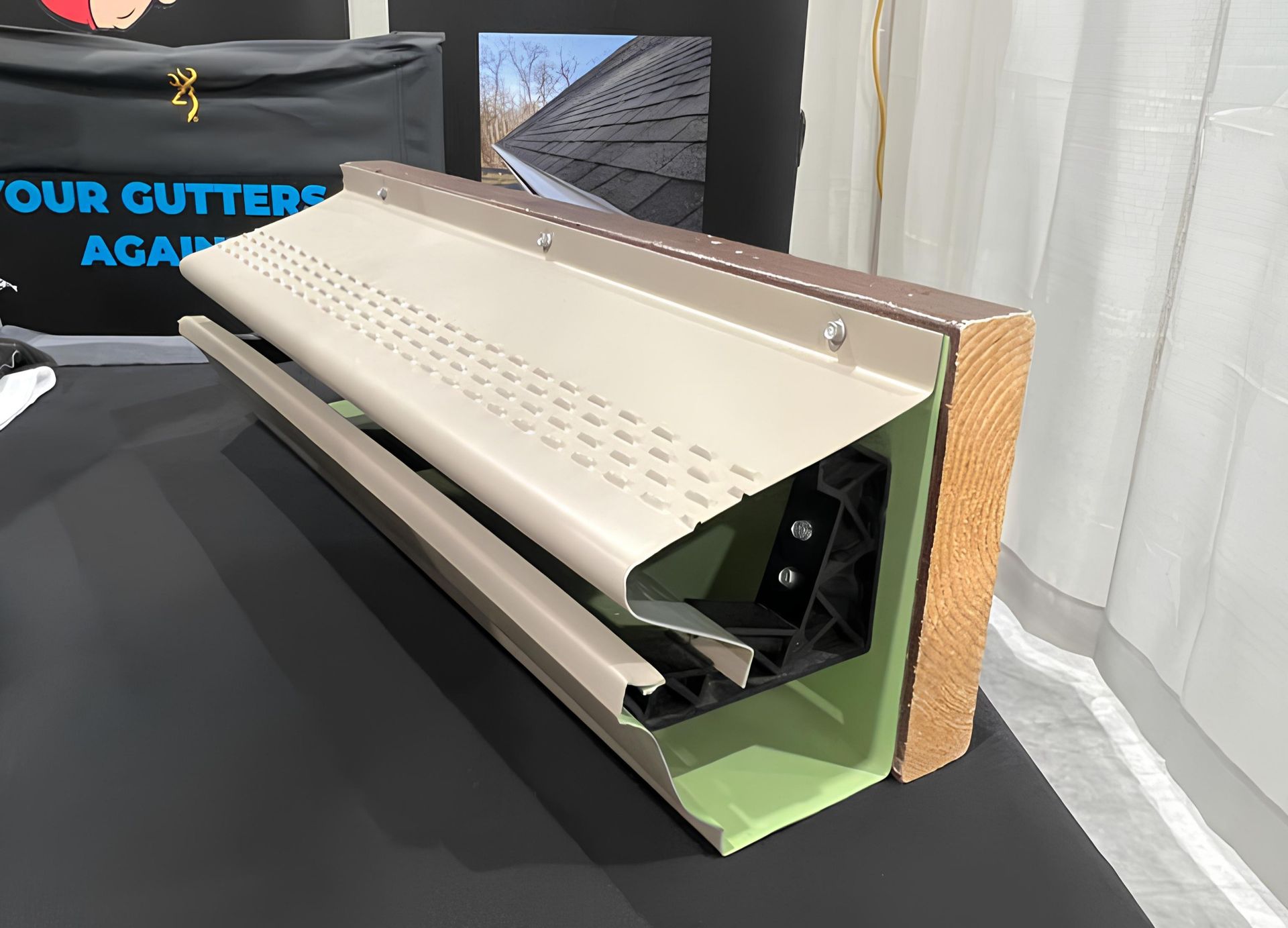 Cutaway view of a gutter system, showing the beige cover, green gutter, and mounting hardware on a wood surface.