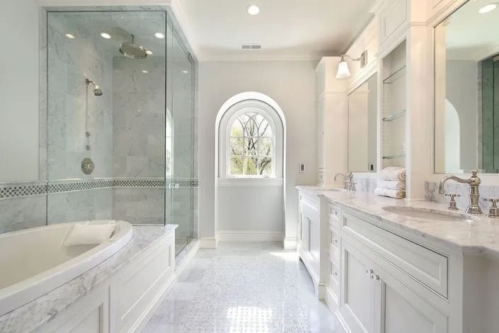 Spacious white bathroom with a glass-enclosed shower, soaking tub, and double vanity. Arched window in the center.