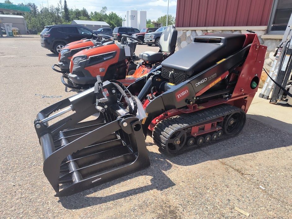 Compact track loader with black grapple attachment parked beside similar equipment outdoors in a lot
