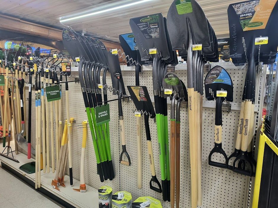 Hardware store aisle with rakes, shovels, and long-handled garden tools hanging on display racks.