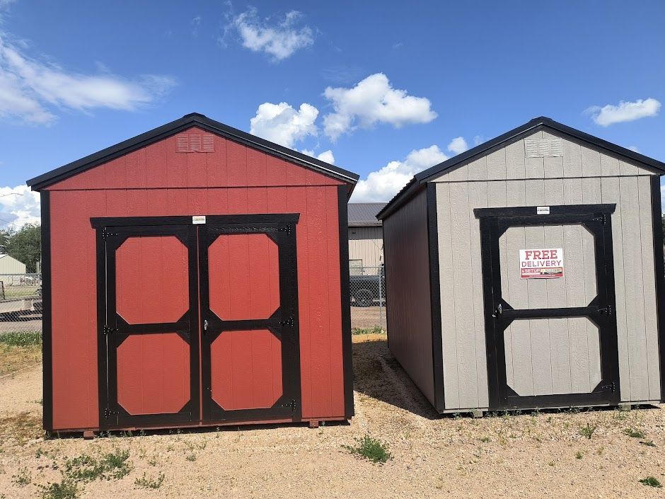 Two small sheds, one red and one white, standing side by side on a gravel lot under a blue sky.