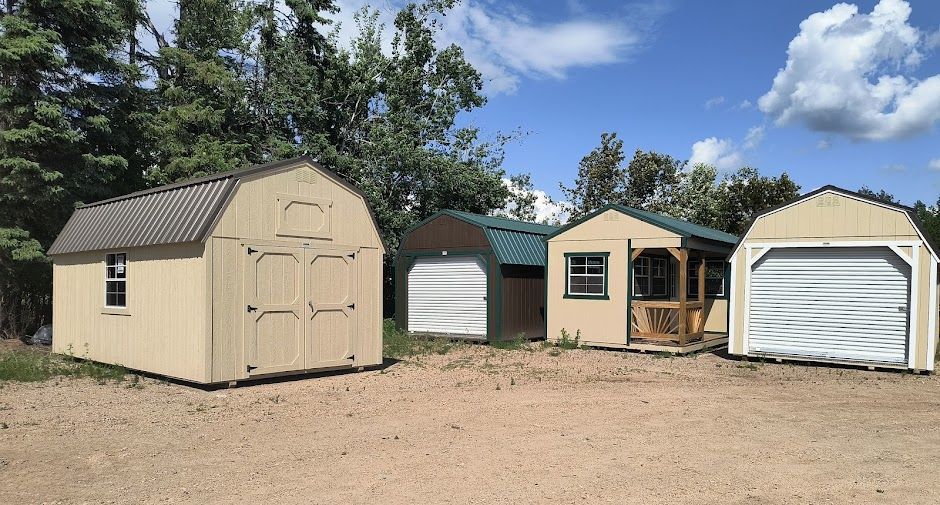 Three beige storage sheds and a white garage-style shed on a dirt lot under a blue sky.