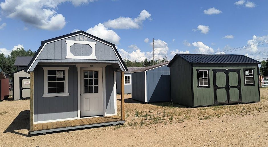 Two small prefabricated cabins, one gray and white and one dark green, on a sandy lot under a blue sky.