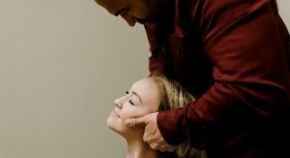 Man in red jacket adjusting woman's neck. Woman looks up with closed eyes, set against plain wall.