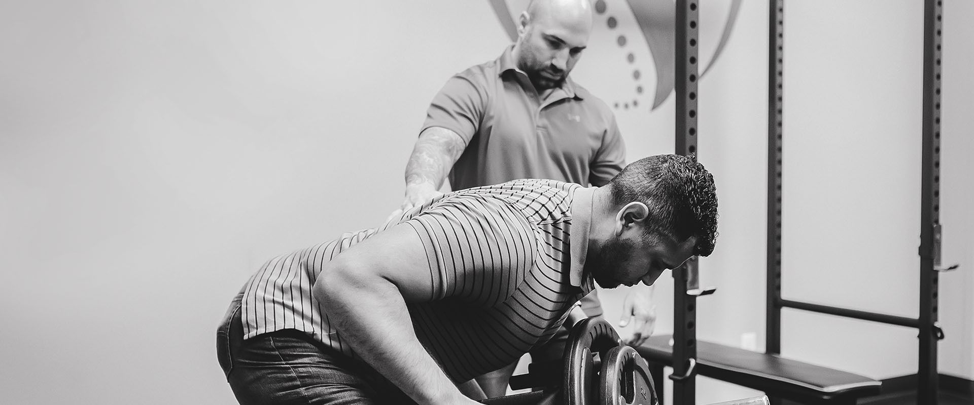 A man assisted by a trainer, bending over while exercising in a gym. Black and white photo.