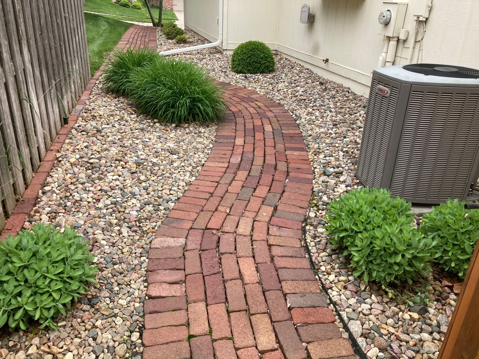 a brick walkway leading to a house with a fence and a air conditioner