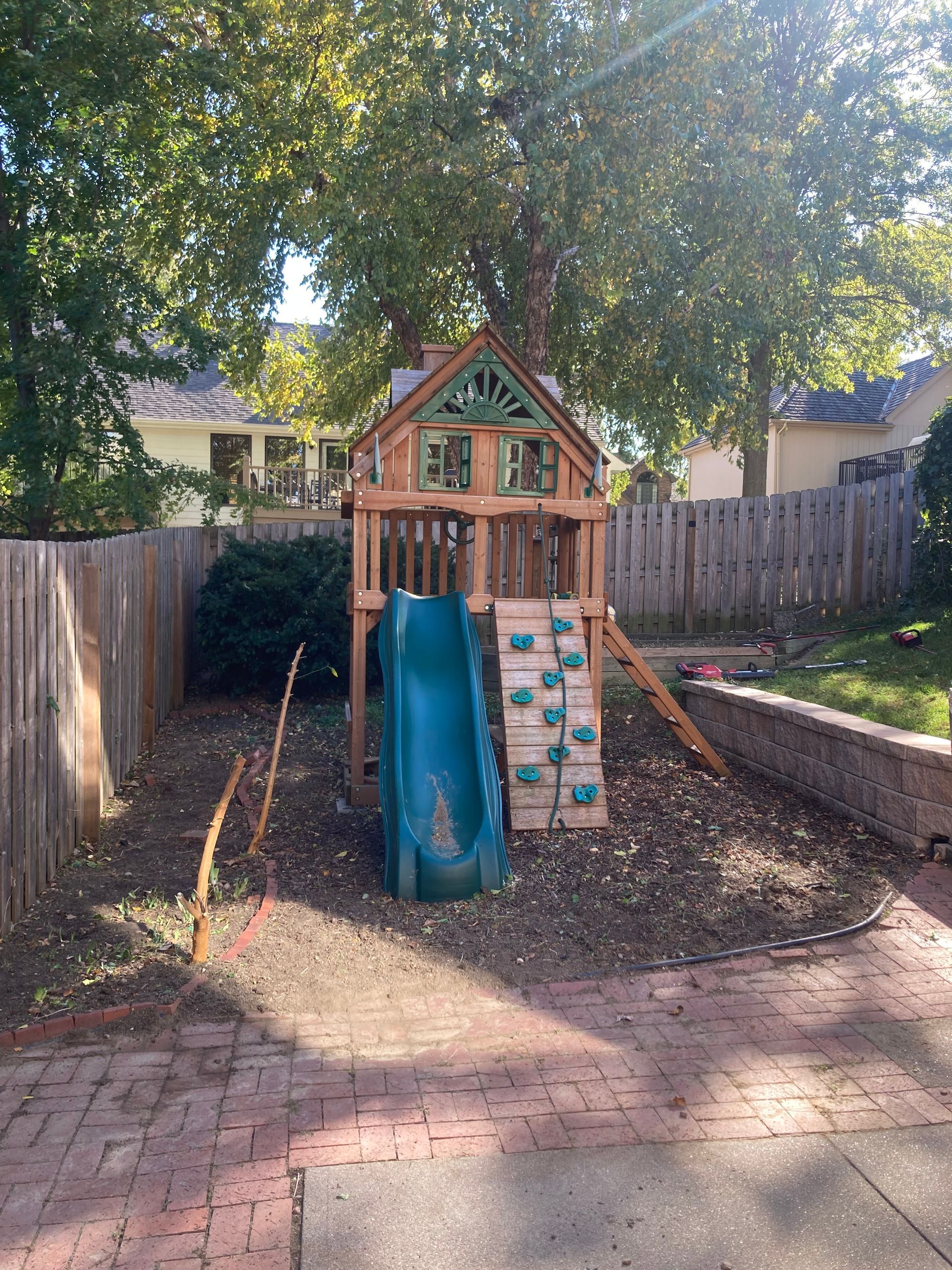 A wooden backyard playset with a green slide and climbing wall sits on mulch behind a brick patio and wood fence.