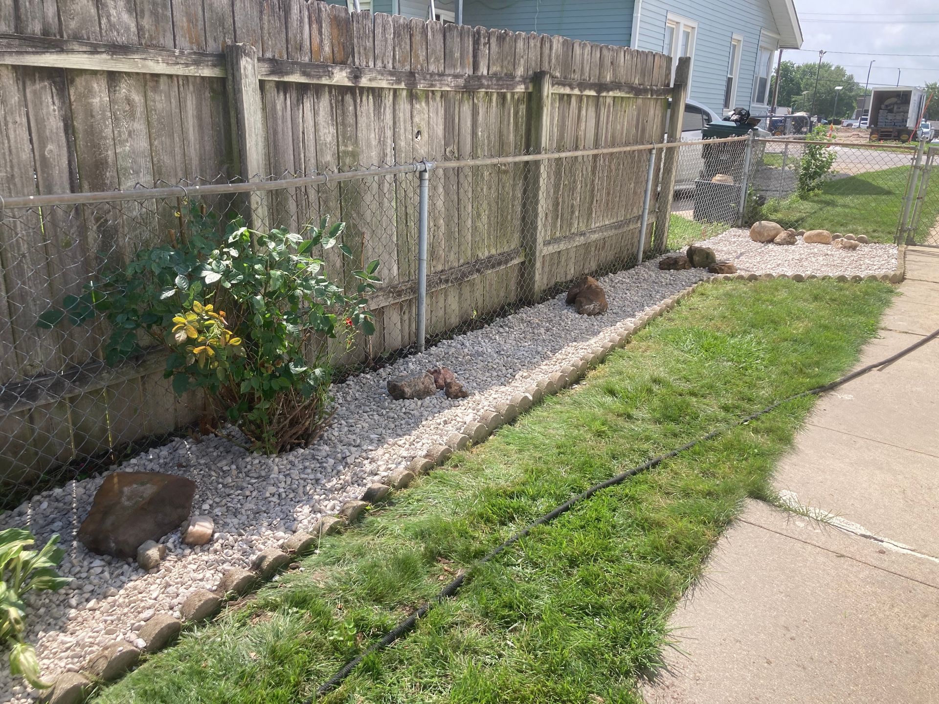 A gravel garden bed runs along a wooden fence, bordered by stones with a small bush and grass lawn in the foreground.