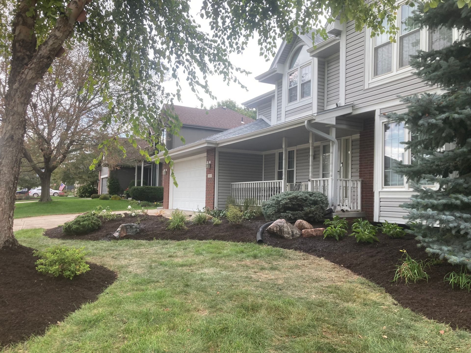 A light gray, two-story house with a front porch, surrounding garden beds covered in dark mulch, and a green lawn.