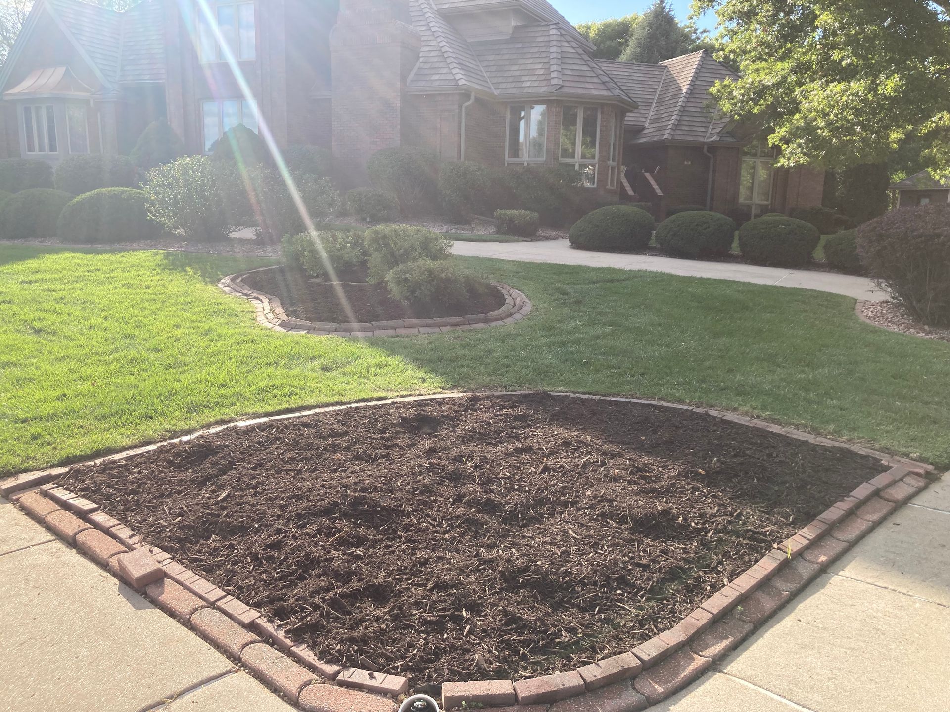 A brick-bordered, mulch-filled garden bed in the foreground, with a lawn and residential home in the background.
