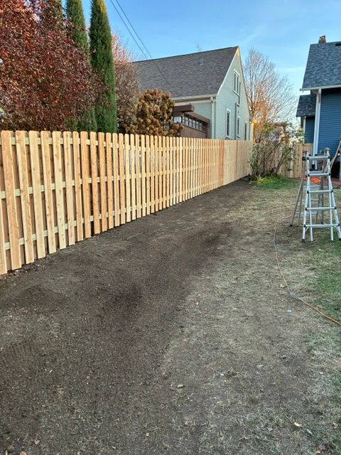 Newly constructed wooden fence bordering a dirt yard, with a ladder leaning against a neighboring house.