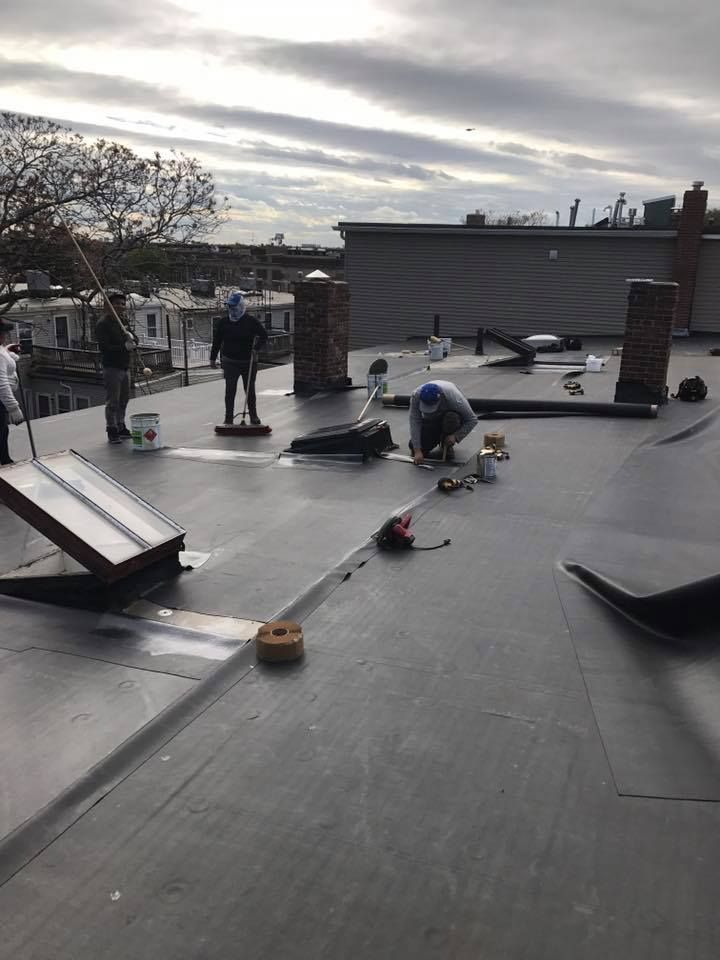 Workers installing a black flat roof on a building; some are bending over, others are standing. Overcast day.