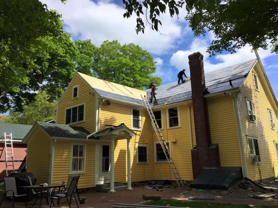 Yellow house with workers on roof installing shingles, ladder in use, blue sky.