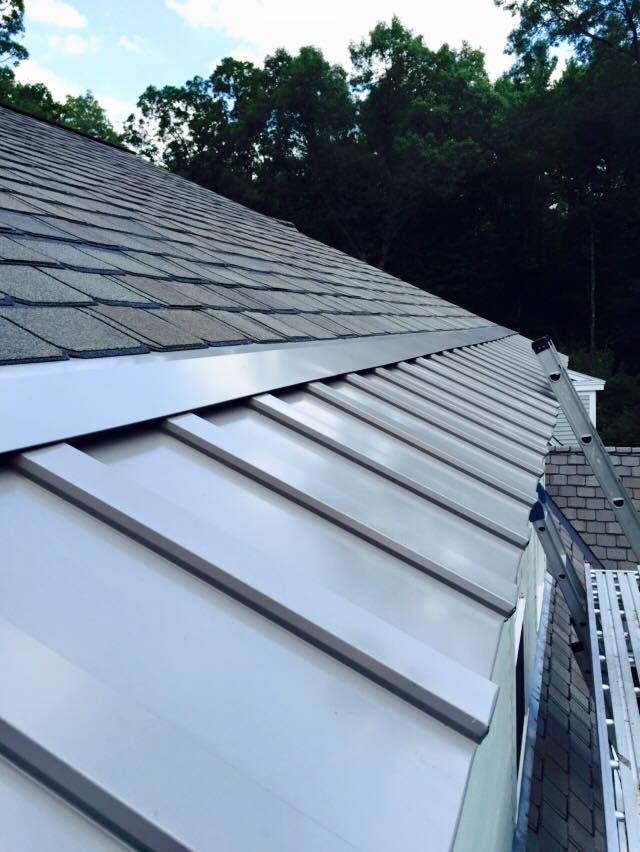 View of a roof with gray asphalt shingles and white metal panels; blue sky and trees in background.