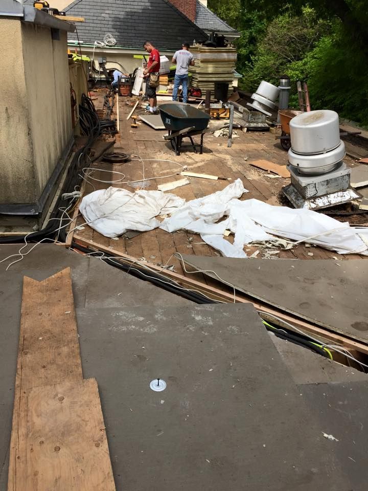 Construction workers on a roof removing old roofing material. Boards, wheelbarrow, and debris are scattered.