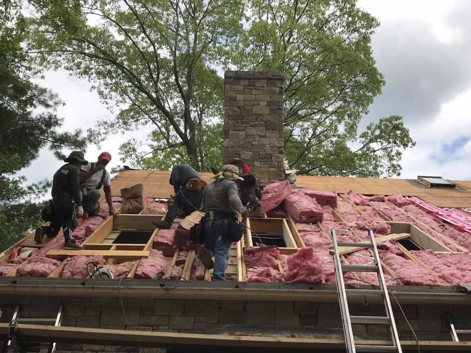 Roofers working on a roof, installing insulation.  Pink insulation and wooden framing visible. A stone chimney stands tall.