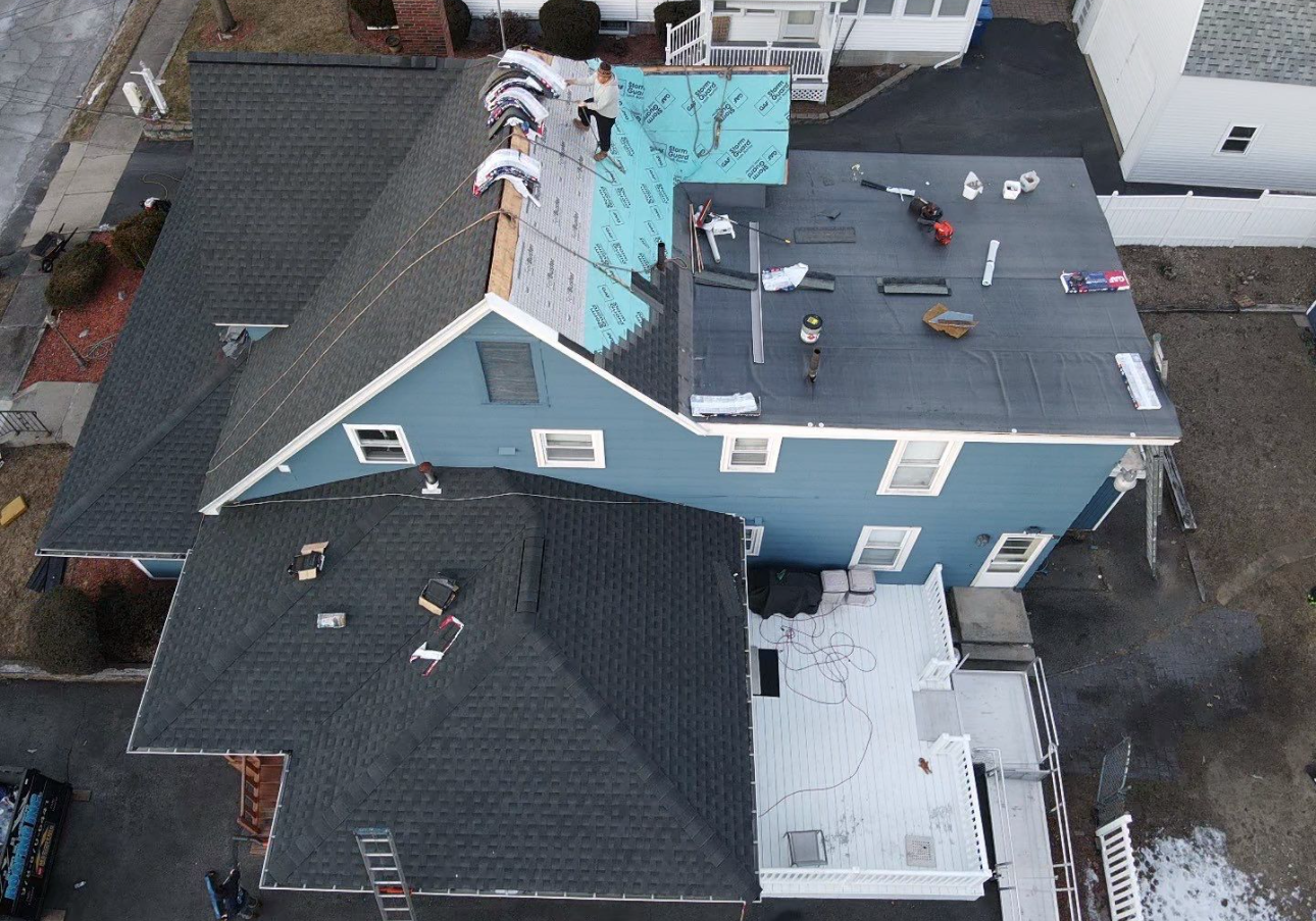 Aerial view of a blue house with roofing in progress; workers on the roof, tools and materials visible.