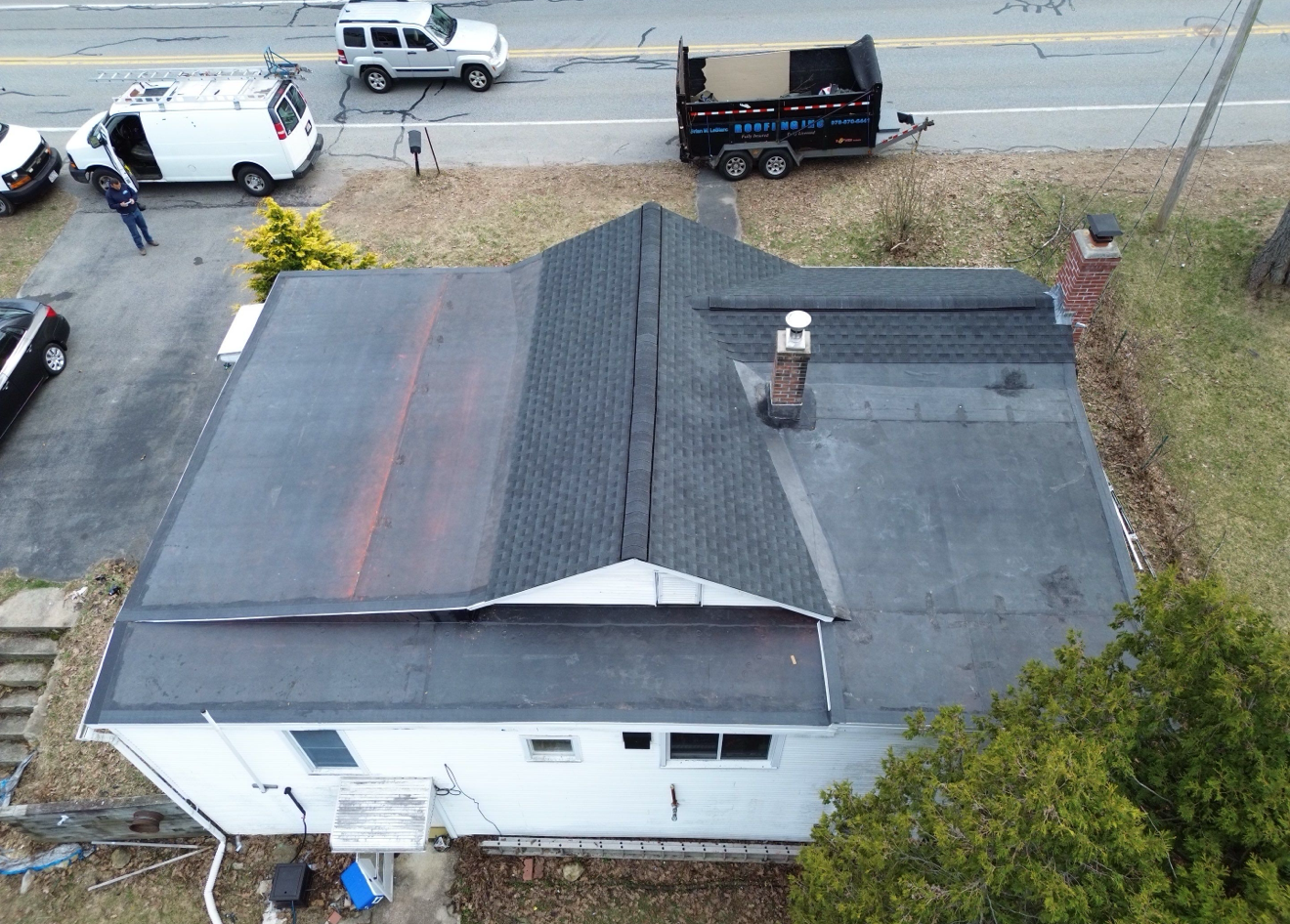 Overhead view of a house with a dark gray roof. A dump trailer, vehicles, and a person are nearby.