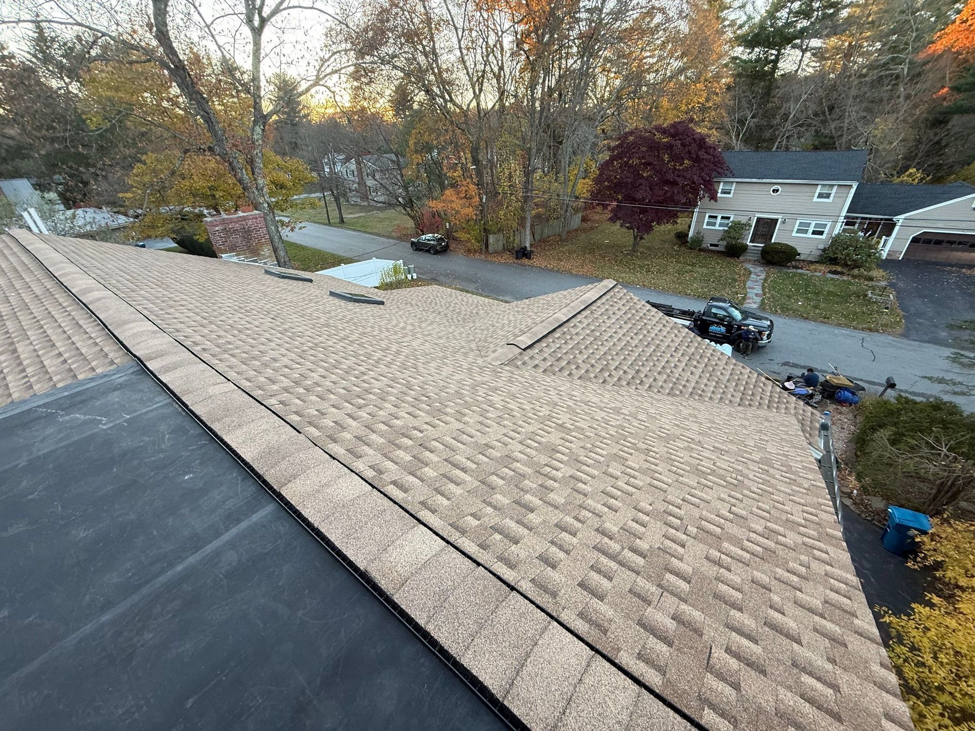 Overhead view of a roof with brown shingles, a black flat section, and a chimney; trees and houses in the background.
