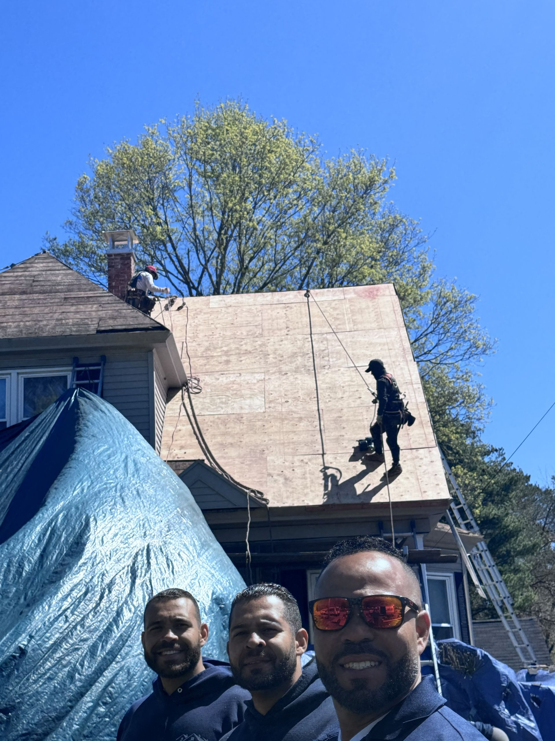Roofing crew installing a new roof on a house, with workers and blue tarp. Sunny day.