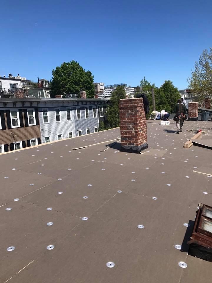 Rooftop with a brick chimney, fasteners, and a worker in the distance, surrounded by buildings on a sunny day.