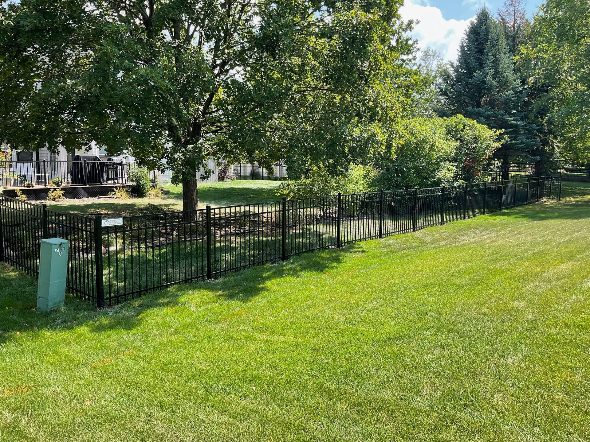 Lush green lawn borders a dark metal fence along a tree-lined area. A green utility box is in the foreground.