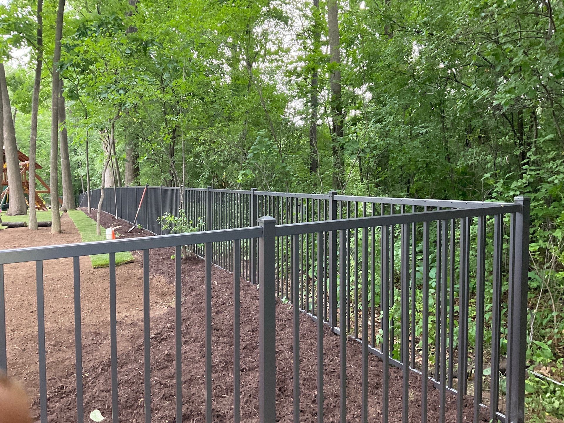 Black metal fence bordering a mulch bed, with a wooded area in the background.