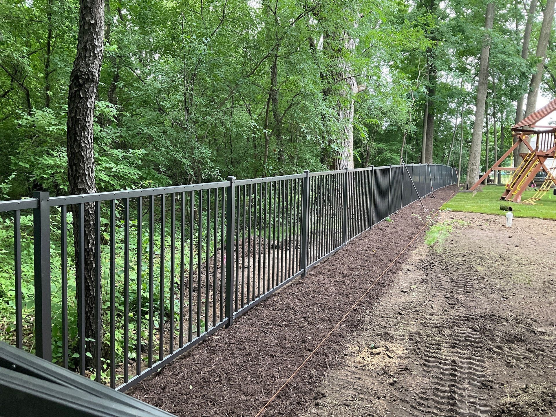 Black metal fence along a wooded area, with a playground visible in the background.