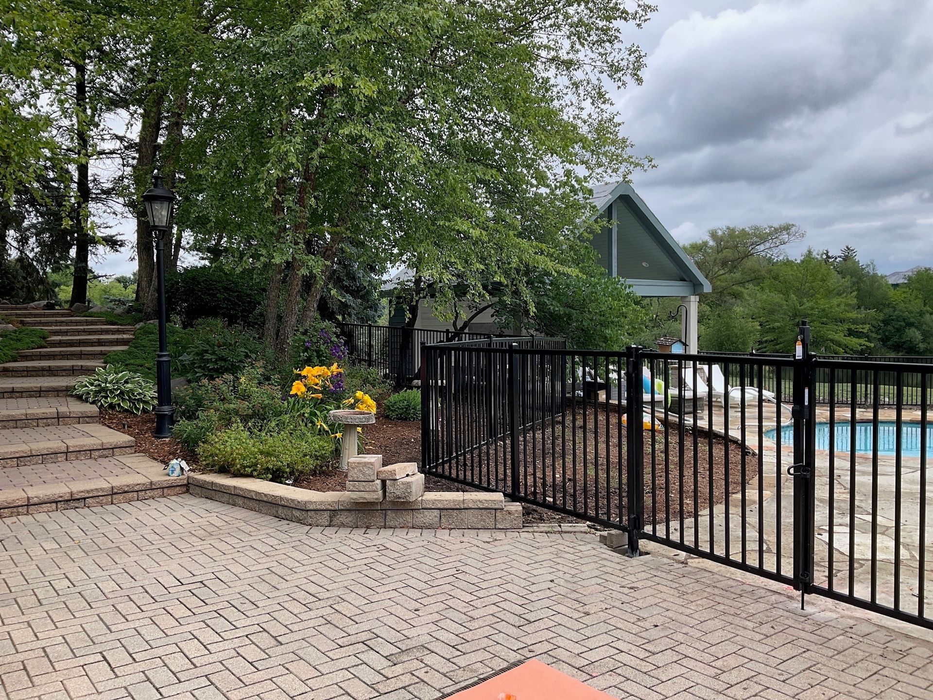 Brick patio next to a black fence bordering a swimming pool and landscaped gardens. Overcast sky.