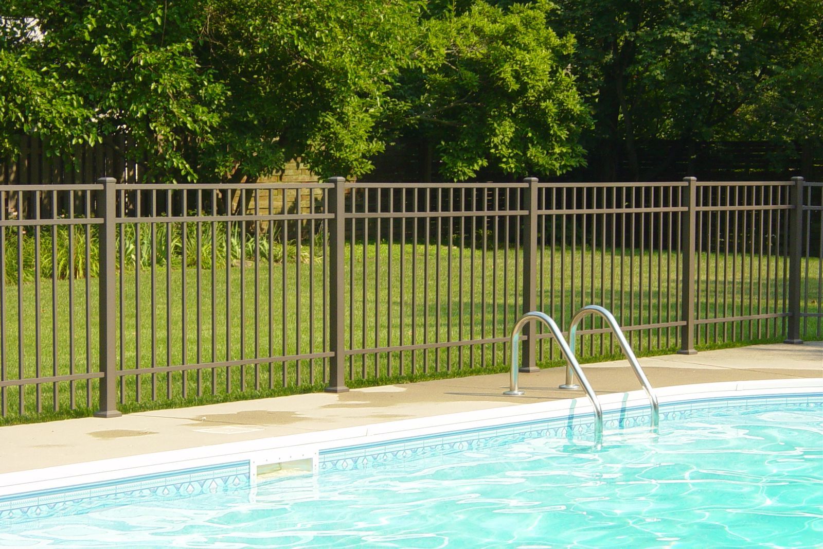 Brown metal fence surrounding a swimming pool with a ladder, green grass, and trees in the background.